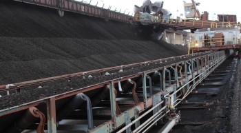 Coal moves down the first of several conveyor belts on its way into the power plant. In the background, a machine scoops the coal down onto the belt from where it was released from the train cars.