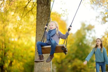 One girl pushes another on a tree swing in a shaded yard.