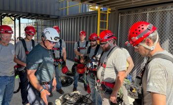 The team gathers, wearing safety helmets and harnesses, to prepare for another challenge. 