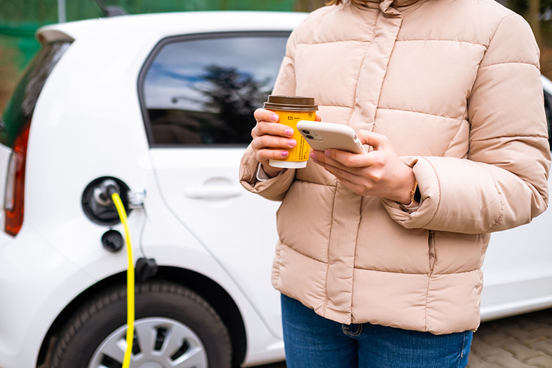 Battery preconditioning can improve your EVs performance, comfort during the winter. In the photo, a woman holds her cellphone and a cup of coffee while standing near her car, which is plugged in.